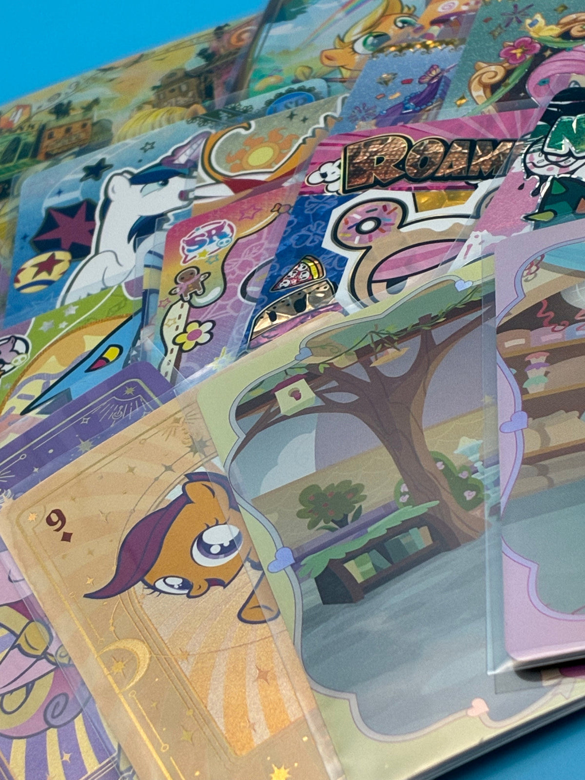 A fanned row of My Little Pony cards in sleeves, pale blue beach and night sky designs behind three gold and pink foil cards.